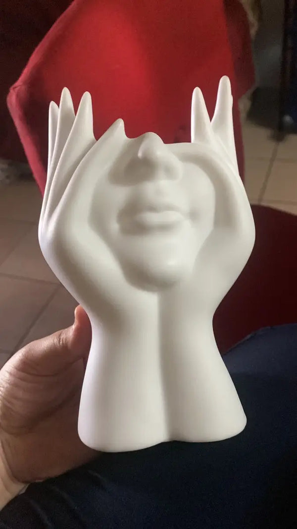 White sculpture of a face held in hands against a red chair background vase auraworks