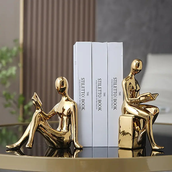 Gold human-shaped bookends on a reflective surface with books in the background. decor aura works