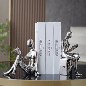 Silver abstract sculptures of people reading books on a surface with a blurred background decor aura works