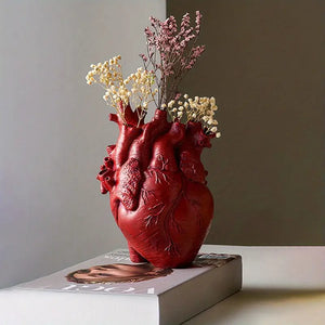 Red heart-shaped vase with flowers on a book against a neutral background