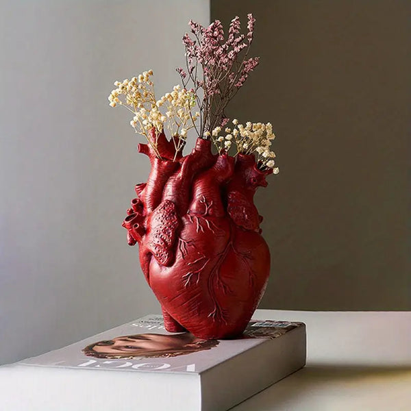Red heart-shaped vase with flowers on a book against a neutral background