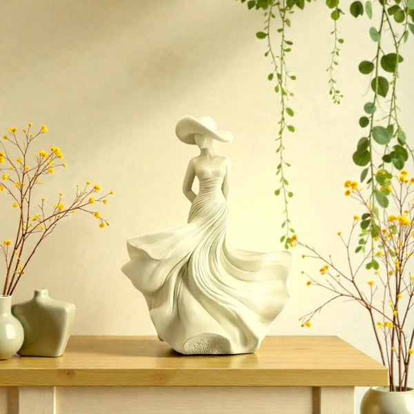 Decorative statue of a woman in a flowing dress on a wooden table with plants and vases. Ivory Serenity Goddess Sculpture