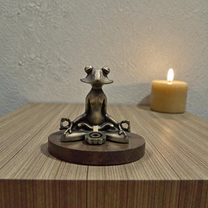 Bronze frog sculpture in a meditative pose on a wooden base with a lit candle in the background.