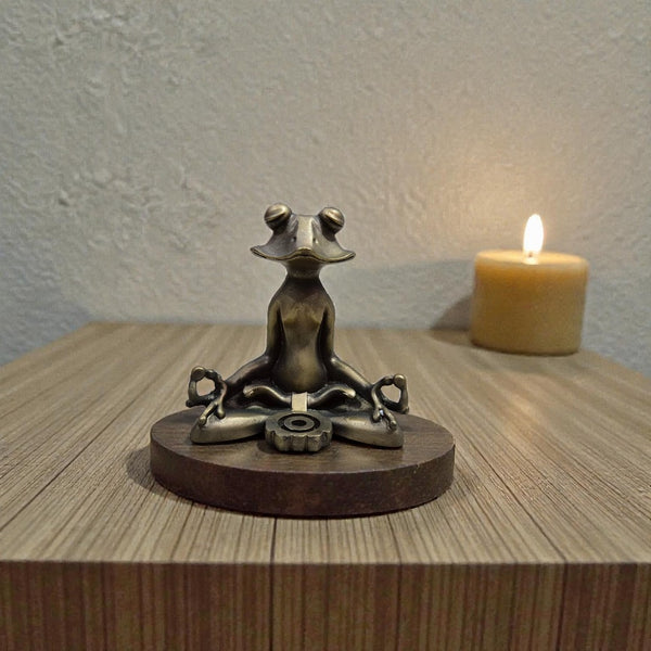 Bronze frog sculpture in a meditative pose on a wooden base with a lit candle in the background.