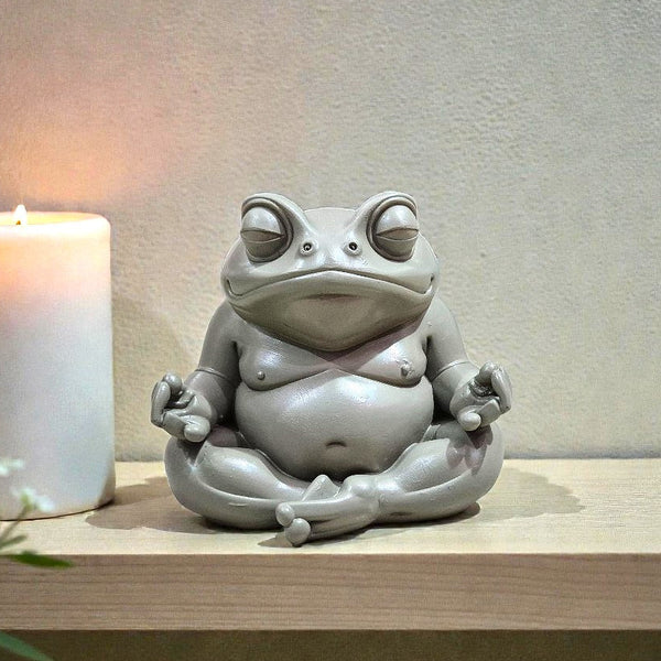 Gray frog sculpture in a meditative pose next to a lit candle on a wooden surface.