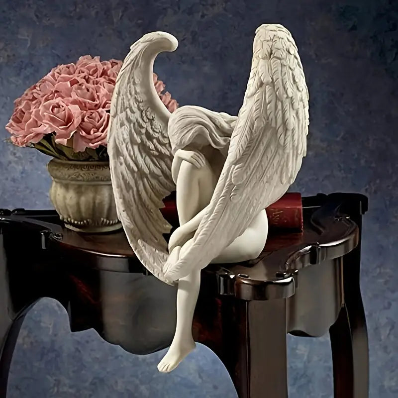 Winged Grace Angel statue in white resin, sitting with folded wings on a wooden table next to a pink rose bouquet – elegant spiritual home decor. Auraworks