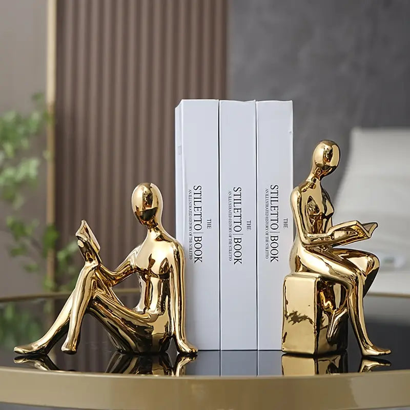 Gold human-shaped bookends on a reflective surface with books in the background. auraworks decor