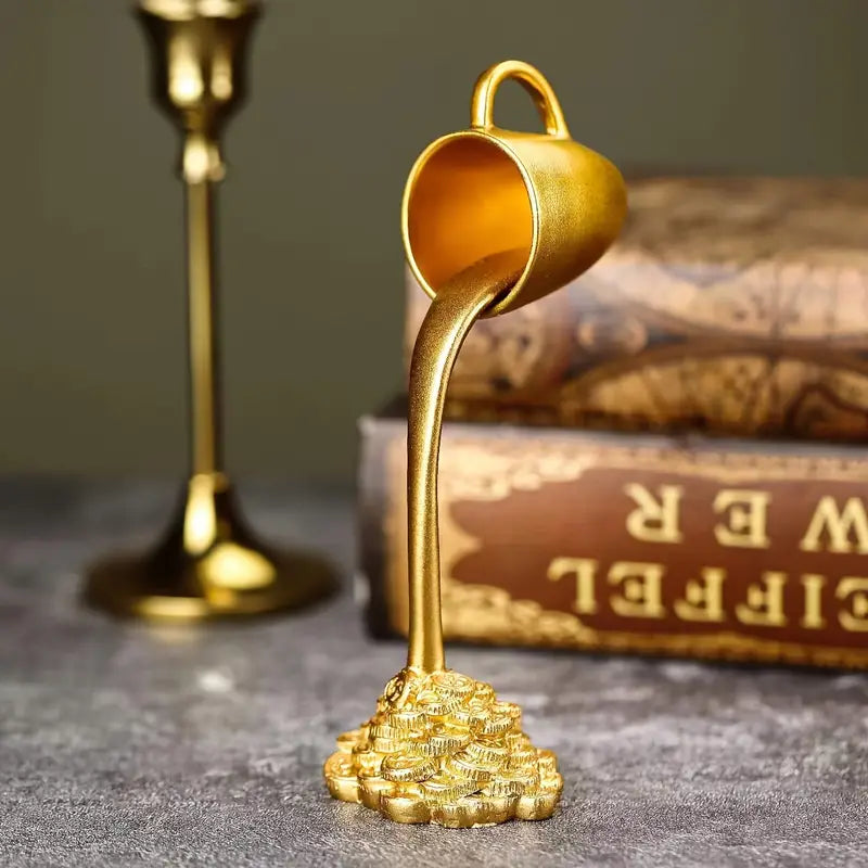 Gold decorative item resembling a teacup pouring gold coins on a textured surface with a book in the background. auraworks decor