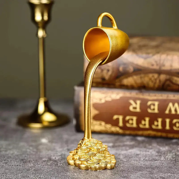 Gold decorative item resembling a teacup pouring gold coins on a textured surface with a book in the background. auraworks decor